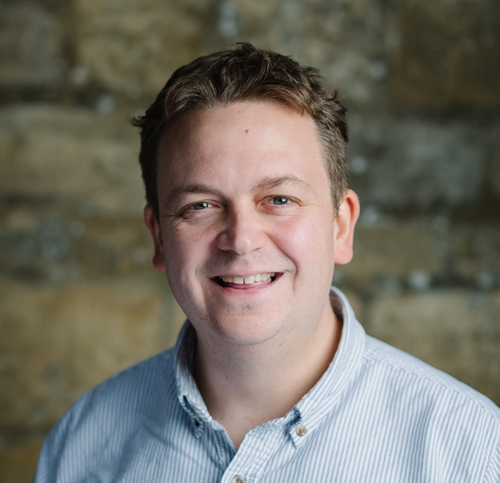 portrait of Tom Knightley smiling against the honey coloured stones of our chapel premises